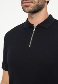Homme portant un polo noir à manches courtes avec une fermeture éclair argentée partiellement fermée au col, laissant apparaître le cou et le bas du visage.
