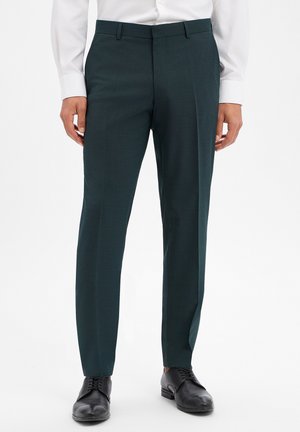 Man draagt donkergroene formele broek, wit overhemd en zwarte leren nette schoenen, staand tegen een witte achtergrond.