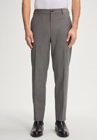 Uomo che indossa pantaloni eleganti grigi su misura e scarpe di pelle nere, in piedi contro uno sfondo chiaro e semplice.