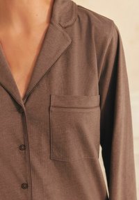 Ruskea pitkähihainen pyjamapaita pehmeästä kankaasta, jossa on viistetty kaulus, napitus edessä ja pieni rintatasku. Sujuva kangas.