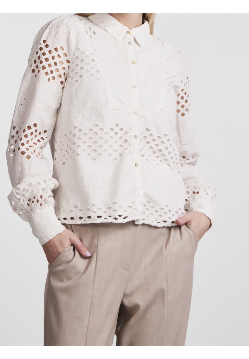 Chemisier blanc boutonné avec motif ajouré en treillis, manches longues, porté rentré dans un pantalon beige taille haute, mains dans les poches.