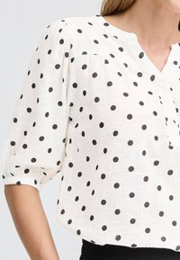 Blouse blanche à pois noirs, manches trois-quarts bouffantes avec poignets élastiques, et patte de boutonnage partielle à l'avant.