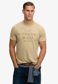 Camiseta de algodón beige con la inscripción "SUPERDRY & CO" en gris oscuro, mangas cortas, cuello redondo, combinada con jeans oscuros y una camisa de cuadros azul atada a la cintura.
