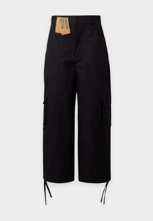Femme debout, mains dans les poches, portant une veste en jean patchwork, une chemise blanche, un pantalon noir large et des chaussures noires pointues sur fond uni.
