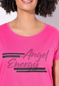Rosa T-shirt med korta ärmar, med svart text "Angel Energy" prydd med rhinstenar och svarta ränder över framsidan.
