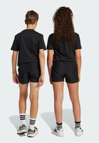 Čierne bavlnené tričká a šortky na dvoch deťoch, stojacich chrbtom k sebe. Čierno-biele pruhované ponožky. Biele a zelené športové topánky.