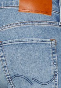 Light blue denim jeans with a brown leather patch at the waist, highlighted stitching, and a rear pocket with curved stitching detail.