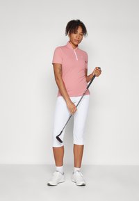 Ροζ πόλο μπλούζα με φερμουάρ στον γιακά, συνδυασμένη με λευκά καπρι παντελόνια και λευκά αθλητικά παπούτσια, κρατώντας ένα μπαστούνι γκολφ.