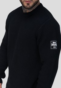 Zwarte geribbelde trui met een ronde halslijn en lange mouwen. Heeft een wit logopatch op de linkerarm. Zachte textuur.