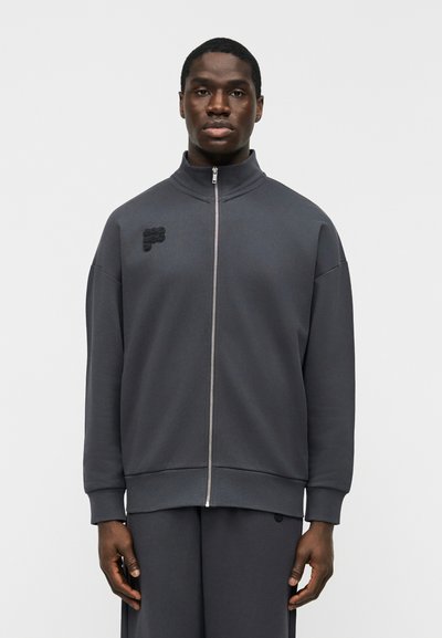 Homme portant une veste zippée gris foncé avec col montant et un pantalon assorti, debout devant un fond clair uni.