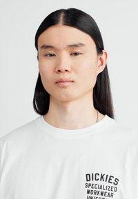 Tricou alb cu grafica textului negru pe piept, decolteu rotund și mâneci scurte. Modelul are părul lung și drept, de culoare neagră.
