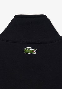 Zwart coltrui kledingstuk met een getextureerde stof en een klein, geborduurd groen krokodillenlogo op de achterkant van de halslijn.