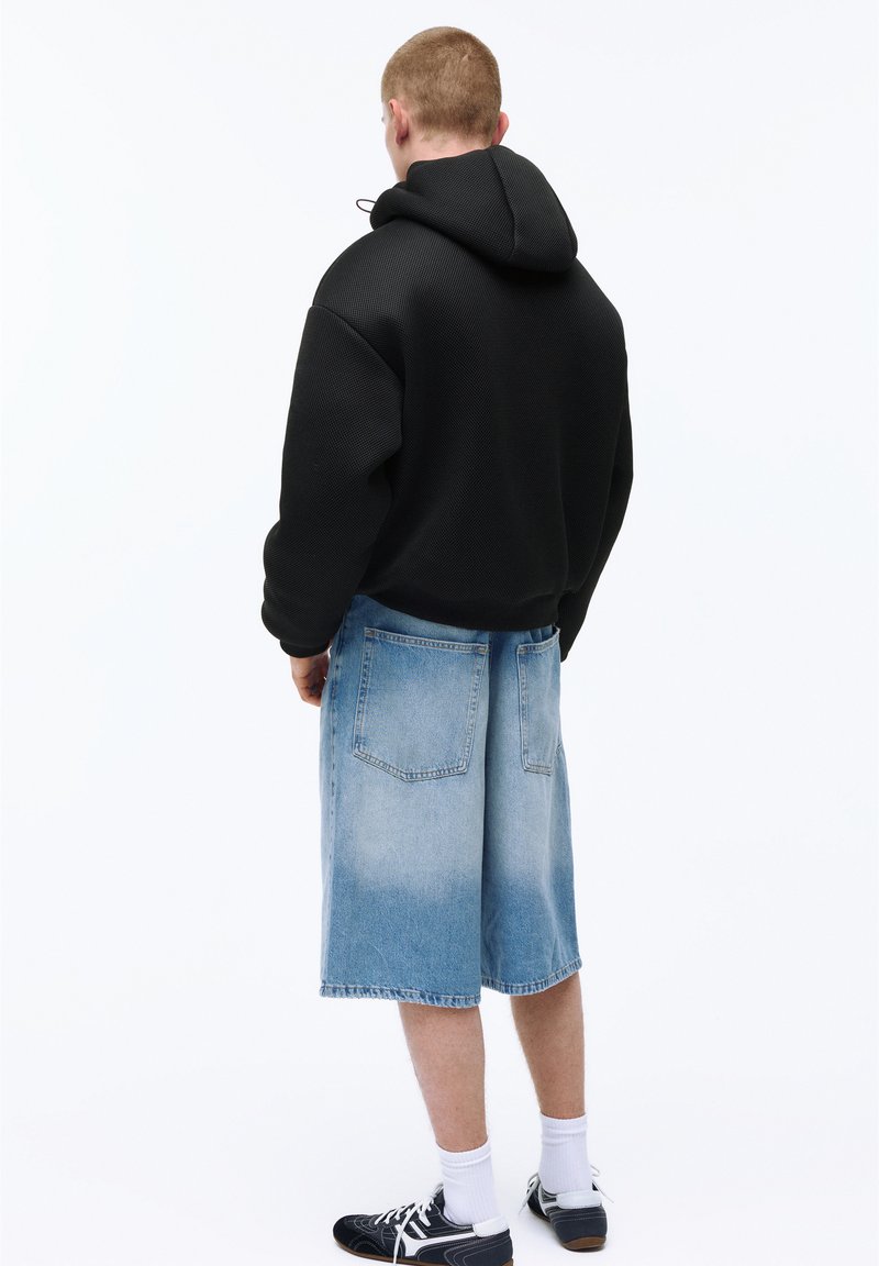 Férfi fekete kapucnis pulóverben, világoskék farmer hosszúnadrágban, fehér zokniban és fekete sportcipőben, háttal áll egy egyszínű háttér előtt.