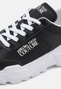 Sapatilha desportiva preta com tecido texturizado e detalhes em pele. Apresenta atacadores brancos, detalhes do logótipo e uma sola branca robusta.