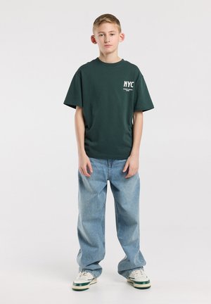 Tienerjongen staand met ontspannen handen, draagt een donkergroen NYC T-shirt, losse lichtblauwe jeans en witte sneakers met groene strepen.