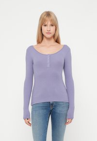 Langermet lilla ribbestrikket topp med rund hals og fem knapper foran. Glatt tekstur, tettsittende fasong. Kombinert med blå jeans.