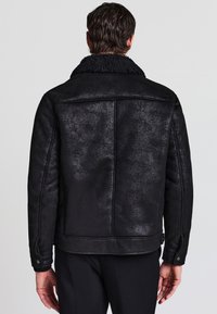 Giacca con colletto in shearling nero, dall'aspetto testurizzato, con una silhouette strutturata, dettagli sui polsini a pressione e linee di cucitura sul retro.