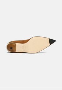 Zapato de tacón alto de ante marrón con punta afilada y(acento) negro en la punta. Suela beige suave con un pequeño corte en la parte trasera.