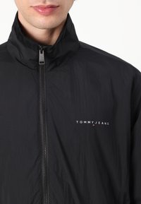 Black lightweight jacket with a high collar, front zipper, and printed "TOMMY JEANS" logo in white on the left side. Smooth texture.