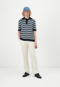 Polo en tricot avec un motif en zigzag bleu, beige et noir, associé à un pantalon crème et des chaussures noires. Casquette en denim sur la tête.