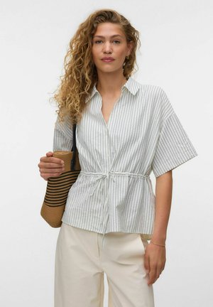 Kvinde med krøllet hår iført en hvid, stribet kortærmet skjorte bundet i taljen, beige bukser og med en brun stribet flettet taske.