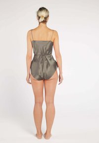 Woman with blonde hair in a bun standing barefoot wearing a gray satin sleeveless romper, facing away against a plain white background.
