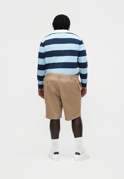 Άτομο που στέκεται με την πλάτη γυρισμένη, φοράει μπλουζάκι με μακριά μανίκια ριγέ σε ανοιχτό και σκούρο μπλε, μπεζ σορτς, λευκές κάλτσες και λευκά αθλητικά παπούτσια.