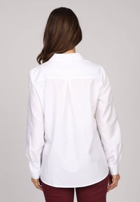 Chemise blanche avec col, manches longues et plis à l'arrière. Tissu lisse et coupe ajustée avec poignets boutonnés.