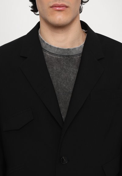 Schwarzer Blazer mit zwei Vordertaschen, Ein-Knopf-Verschluss und einer strukturierten grauen Innenschicht, die am Kragen sichtbar ist. Glatte Stoffoberfläche.