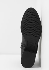 Zapato de cuero negro con una suela de goma texturizada que presenta surcos y orificios de ventilación. Costuras laterales y detalle de logo en relieve visibles.