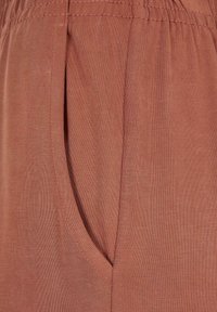 Gros plan d'un pantalon en tissu de couleur rouille avec une taille élastique et une poche latérale visible à gauche.
