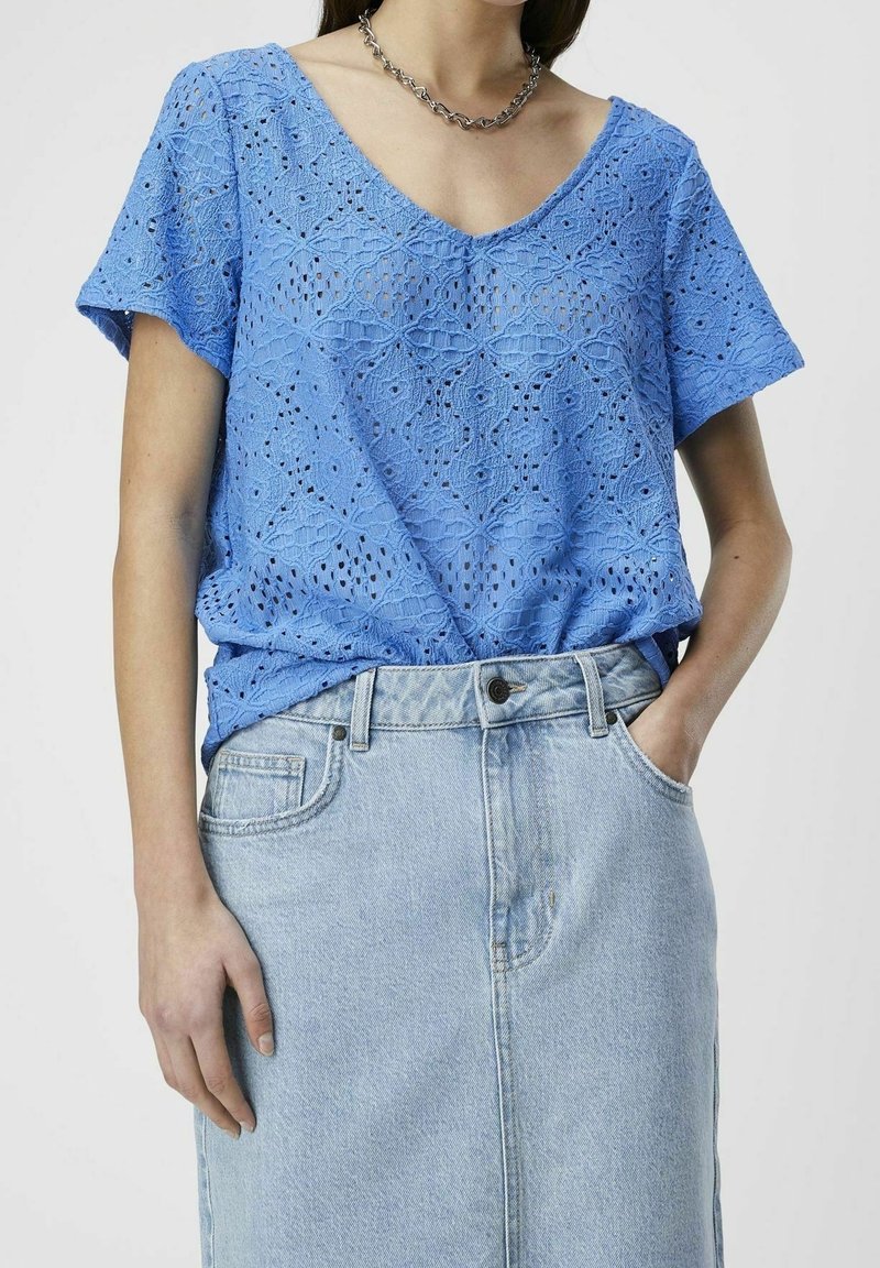 Blause aus blauer Baumwollspitze mit einem V-Ausschnitt, kurzen Ärmeln und einem strukturierten Muster, getragen über einem hellen Denim-Rock mit Knopf- und Taschendetails.
