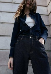 Camicia in denim scuro con chiusura a bottoni e maniche lunghe abbinata a jeans scuri a vita alta. Entrambi presentano cuciture a contrasto e tasche.