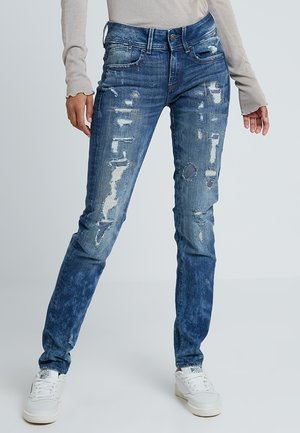 Jeans azules desgastados con tonos variados, que presentan desgarros y parches, diseño de cinco bolsillos y una silueta ajustada.