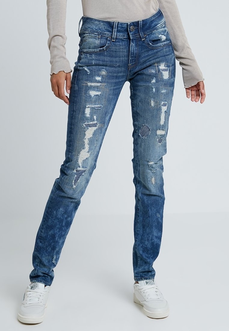 Jeans azules desgastados con tonos variados, que presentan desgarros y parches, diseño de cinco bolsillos y una silueta ajustada.