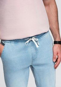 Světle modré džínové joggers s elastickým pasem, bílou šňůrkou, bočními kapsami a měkkou strukturou. Nosené s růžovým topem.