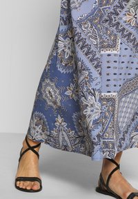 Robe maxi à motif bleu avec des designs paisley en tons blancs et bruns, assortie à des sandales noires à lanières. Tissu doux et forme fluide.