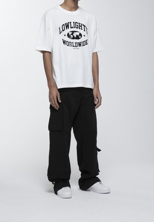 Camiseta de algodón blanca con estampado gráfico negro que dice "LOWLIGHTS WORLDWIDE", combinada con pantalones cargo negros holgados y zapatillas blancas.