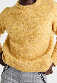 Gul texturerad tröja med rund halsringning och långa ärmar, kombinerat med rutiga byxor som har dragkedjor på sidorna.