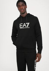 Felpa nera con tasca a marsupio frontale, cappuccio con cordoncino e logo EA7 Emporio Armani bianco. Caratterizzata da polsini e orlo a coste.