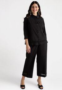 Svart lagerblus med knäppning fram, trekvartslånga ärmar och spetsdekoration vid nederkanten, i kombination med vida svarta byxor och klackar.