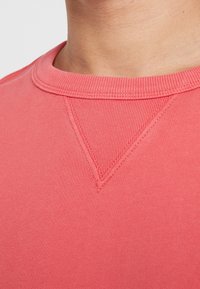 Koraalkleurige sweatshirt met een ronde halslijn, ribgebreide kraag en driehoekige stikdetails op de schouder. Zachte stoftextuur.