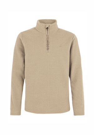 Junge trägt beigen Fleecepullover, dunkelblaue Schneehose, schwarze Stiefel und dunkelblaue Strickmütze und steht vor einem schlichten hellen Hintergrund.