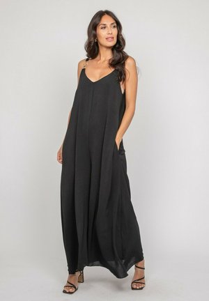 Femme portant une robe maxi noire sans manches avec des bretelles réglables et des poches, debout devant un fond uni clair.