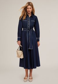 Rochie de stil cămașă, de culoare bleu marin, cu mâneci lungi, accente albe, talie strânsă și fustă amplă. Asortată cu o geantă mică bej și pantofi cu vârful ascuțit.