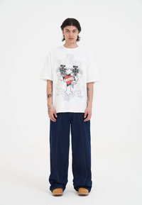 T-shirt bianco oversize con grafica di croci e testo. Abbinato a pantaloni in denim scuri a gamba larga e sandali marroni.