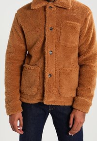 Veste en polaire marron avec un devant à boutons, deux poches carrées et un col. Texture douce avec une coupe légèrement surdimensionnée.