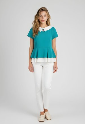 Joven mujer con blusa peplum color azul verdoso con dobladillo y cuello blanco en capas, combinada con pantalones blancos y mocasines beige, de pie sobre un fondo gris.