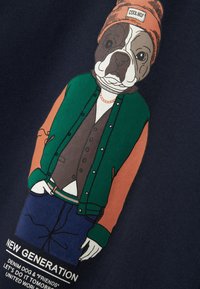 Koszulka z grafiką przedstawiającą buldoga w zielonej i pomarańczowej kurtce typu varsity, beżowej kamizelce i niebieskich dżinsach; napis brzmi "NOWA POKOLENIE."