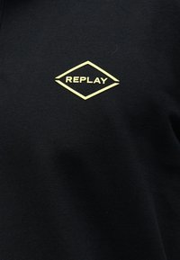 Μαύρο ύφασμα με ένα μικρό κίτρινο λογότυπο σε σχήμα ρόμβου με τη λέξη "REPLAY" στο κέντρο της επιφάνειας.
